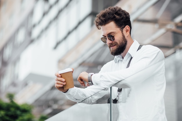 Free photo a bearded serious stylish manager looking to his watch on the streets of the city near modern office centre man drinks coffee an employee looks at the time