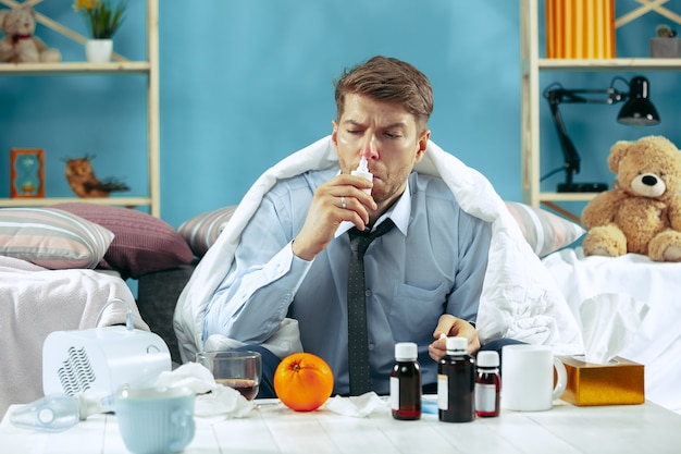 Free photo bearded sick man with flue sitting on sofa at home covered with warm blanket and using nasal spray. the illness, influenza, pain concept. relaxation at home