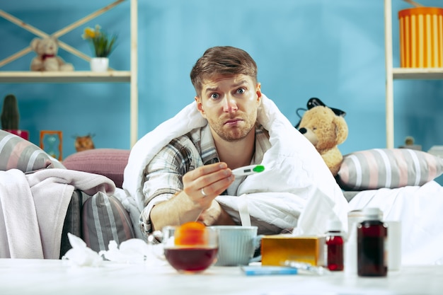 Free photo bearded sick man with flue sitting on sofa at home and measuring body temperature. the winter, illness, influenza, pain concept. relaxation at home