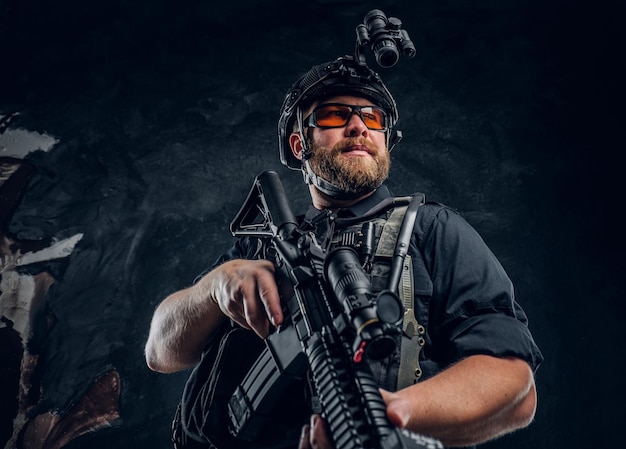 Free photo bearded special forces soldier wearing body armor and helmet with night vision holding an assault rifle. studio photo against a dark textured wall