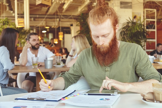 Free Photo bearded student working on course paper, making research, analyzing statistics on tablet, writing down in notebook using pencil.