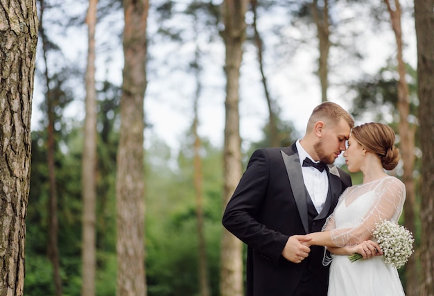 A bearded, stylish groom in a suit and a beautiful blonde bride in a white dress with a bouquet in her hands are standing and hugging in nature in the pine forest.