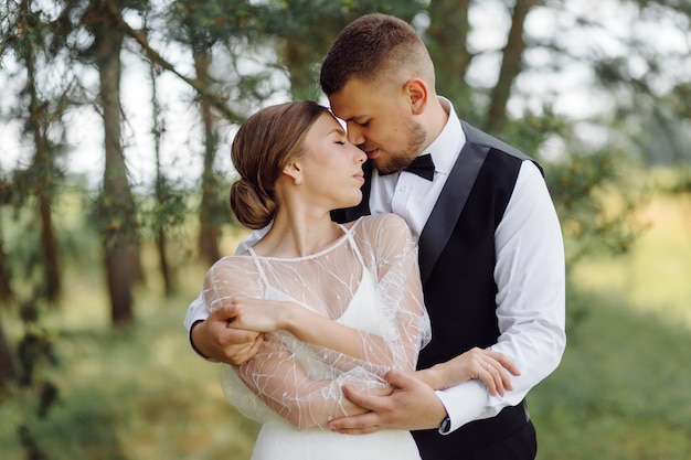 A bearded, stylish groom in a suit and a beautiful blonde bride in a white dress with a bouquet in her hands are standing and hugging in nature in the pine forest.