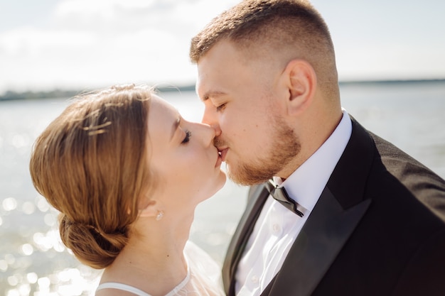 Free photo a bearded, stylish groom in a suit and a beautiful blonde bride in a white dress with a bouquet in her hands are standing and hugging in nature in the pine forest.