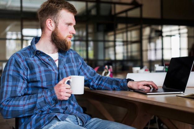 Free photo bearded young businessman working on laptop with holding coffee cup