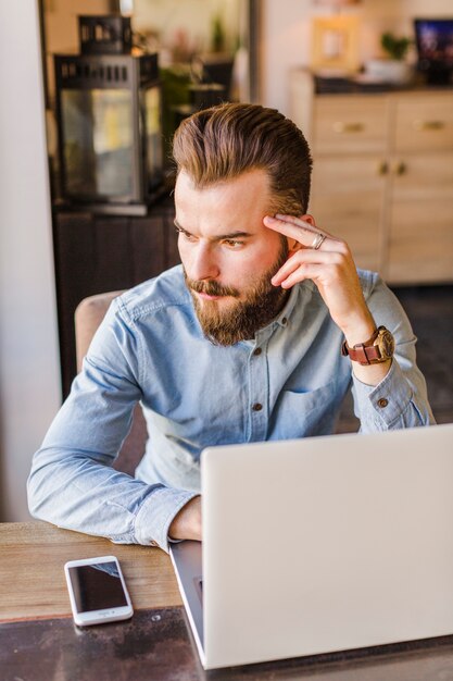 Bearded young man sitting in restaurant with laptop and cellphone on desk