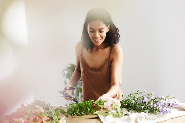 Free photo beautiful african female florist smiling cutting stems working in flower shop over white wall.