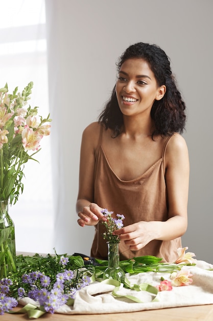 Beautiful african woman florist smiling making bouquet in flower shop over white wall.