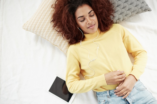 Free photo beautiful african woman in headphones listening to music lying on bed smiling with