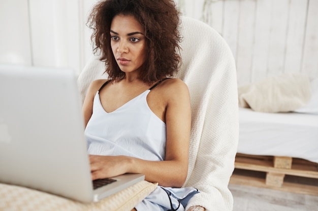 Beautiful african woman in sleepwear looking at laptop sitting in chair at home. 