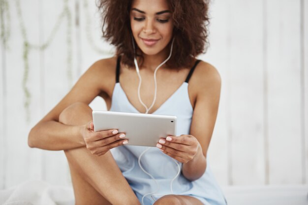 Beautiful african woman in sleepwear smiling looking at tablet listening to music in headphones sitting on bed.