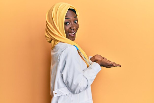 Beautiful african young woman wearing doctor uniform and hijab pointing aside with hands open palms showing copy space presenting advertisement smiling excited happy