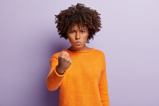 Free photo beautiful annoyed woman raises fist, has frustrated and furious outraged expression, expresses aggression, rage, dressed in orange jumper, isolated over purple wall, promises to revenge.