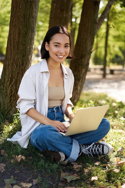 Beautiful asian girl sitting in park with laptop working on remote typing on keyboard smiling at