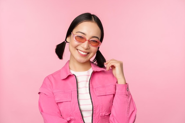 Beautiful asian girl in sunlgasses smiling at camera posing against pink studio background