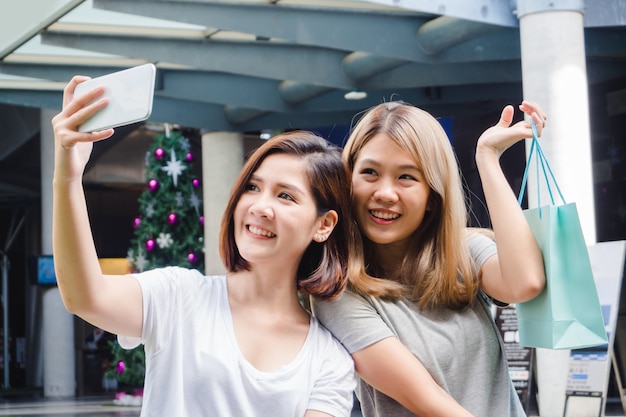 Free photo beautiful asian girls holding shopping bags, using a smart phone selfie and smiling