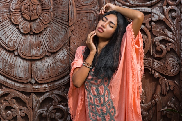 Beautiful asian model in pink boho dress posing over wood ornamental wall.