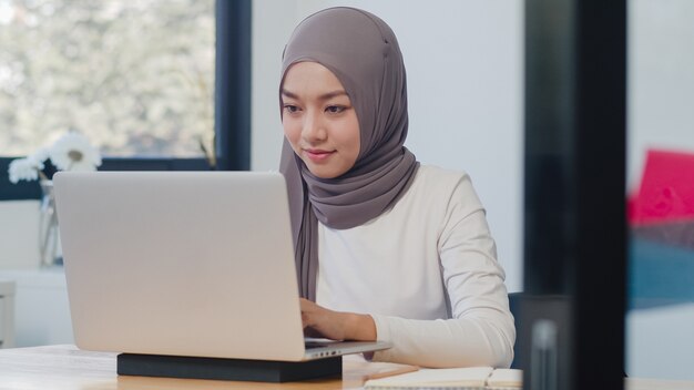 Beautiful Asian muslim lady casual wear working using laptop in modern new normal office.