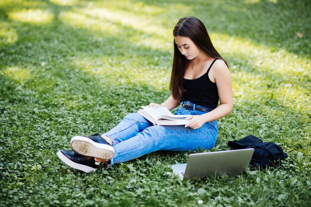 Beautiful asian student keeping records in paper notebook while studying online with laptop, sitting on grass, outdoor