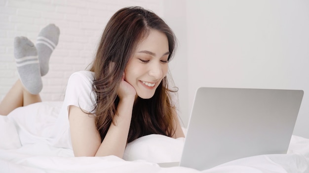 Beautiful Asian woman playing computer or laptop while lying on the bed in her bedroom.