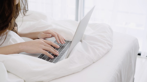 Beautiful Asian woman playing computer or laptop while lying on the bed in her bedroom