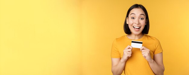 Beautiful asian woman showing credit card and smiling recommending bank service standing over yellow background