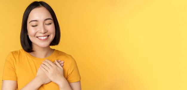 Beautiful asian woman smiling with tenderness and care holding hands on heart standing in tshirt ove