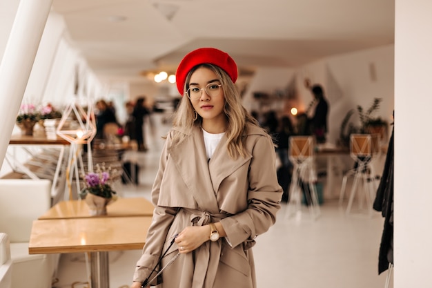 Beautiful Asian woman in trendy trench coat and bright beret holds bag and looks into front against cozy room