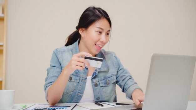 Beautiful Asian woman using laptop buying online shopping by credit card while wear casual sitting on desk in living room at home. 