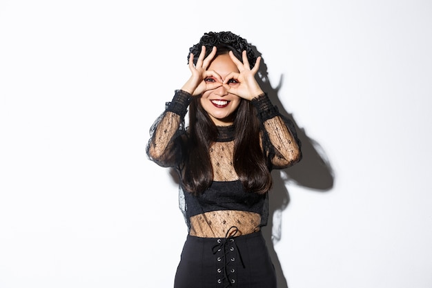 Beautiful asian woman in witch costume looking through finger glasses and smiling happy, standing over white background.