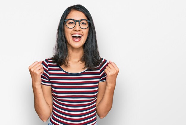 Beautiful asian young woman wearing casual clothes and glasses celebrating surprised and amazed for success with arms raised and open eyes winner concept