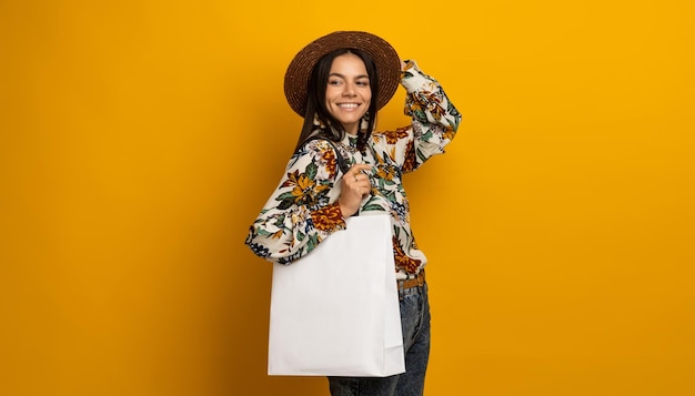 Beautiful attractive stylish brunette woman posing isolated on yellow studio background in trendy fashion clothes outfit holding white shopping bag printed blouse and hat smiling happy accessories