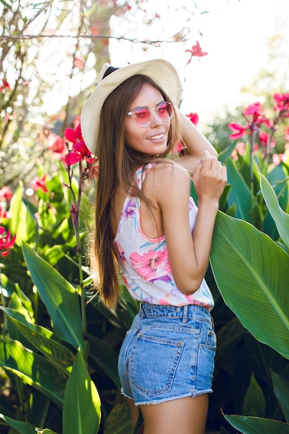 Beautiful beach girl standing in a park wearing straw hat, pink eyeglasses, denim shorts and bright t-shirt. Girl is very slim and attractive. She looks playful. She stands among tropical flowers