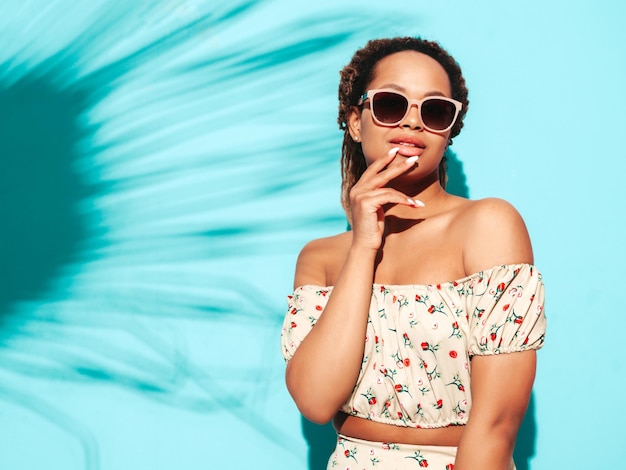 Beautiful black woman with afro curls hairstyle Smiling model dressed in summer hipster clothes Sexy carefree female posing near blue wall in studio Tanned and cheerful In sunglasses