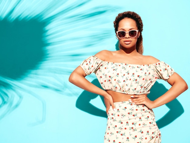 Free photo beautiful black woman with afro curls hairstyle smiling model dressed in summer hipster clothes sexy carefree female posing near blue wall in studio tanned and cheerful in sunglasses