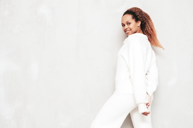 Free Photo beautiful black woman with afro curls hairstyle smiling model dressed in white summer costume sexy carefree female posing near grey wall in studio tanned and cheerful shocked and surprised