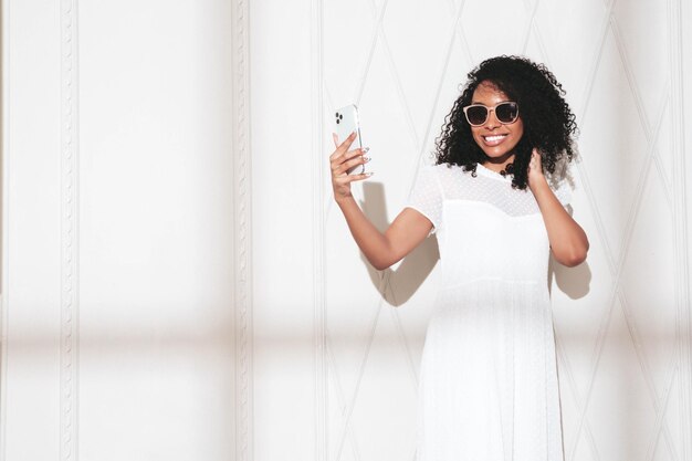 Beautiful black woman with afro curls hairstyle Smiling model dressed in white summer dress Sexy carefree female posing near wall in studioAt sunny day Shadow from window Taking selfie in eyewear