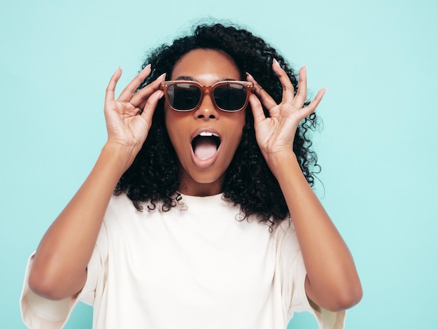 Beautiful black woman with afro curls hairstyle Smiling model in long tshirt clothes Sexy carefree female posing near blue wall in studio Tanned and cheerful in sunglassesShocked and surprised