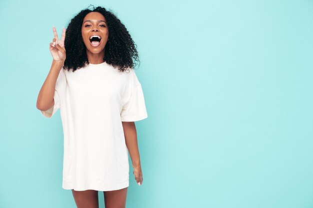 Beautiful black woman with afro curls hairstyle Smiling model in long tshirt clothes Sexy carefree female posing near blue wall in studio Tanned and cheerful