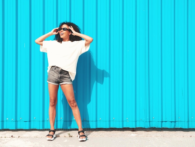 Free photo beautiful black woman with afro curls hairstylesmiling hipster model in white tshirt sexy carefree female posing in the street near blue wall in sunglasses cheerful and happy