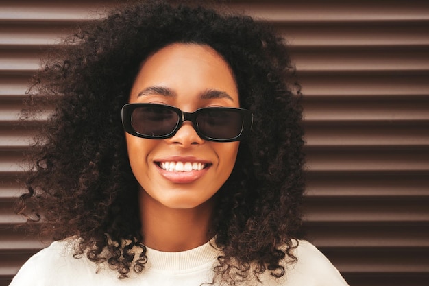 Beautiful black woman with afro curls hairstyleSmiling hipster model in white tshirt Sexy carefree female posing in the street near brown wall Cheerful and happy in sunglasses