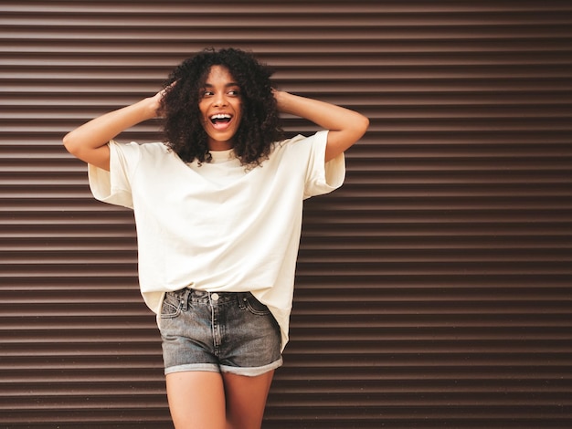 Beautiful black woman with afro curls hairstyleSmiling hipster model in white tshirt Sexy carefree female posing in the street near brown wall Cheerful and happy in sunglasses