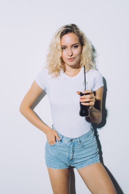 Free photo beautiful blonde girl drinking cola from bottle in casual summer clothes on white wall