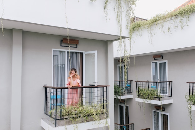 Beautiful blonde girl posing at hotel balcony. smiling romantic woman in pink dress enjoying morning.