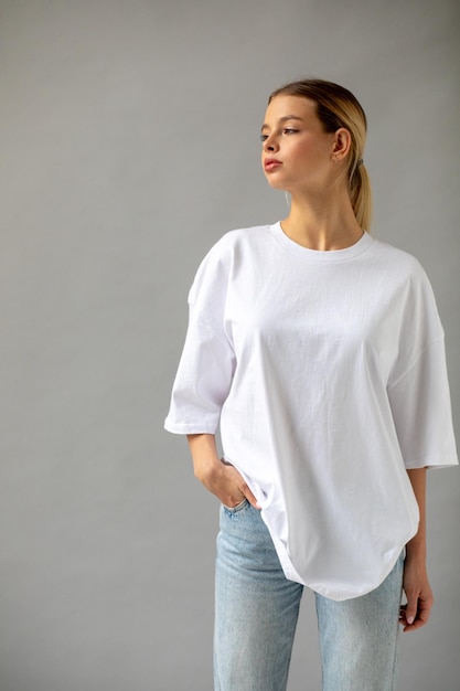 Free Photo beautiful blonde girl in a white oversized tshirt and blue jeans posing on a gray background