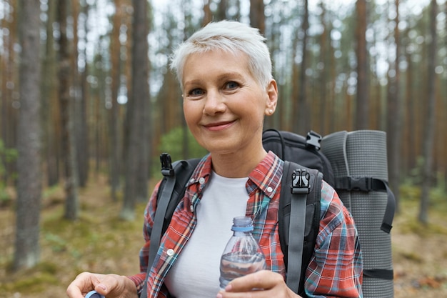 Beautiful blonde retired female spending vacations outdoors, walking in forest with rucksack behind her back, holding plastic bottle of water