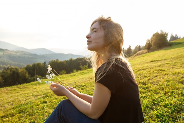 Free photo beautiful blonde woman enjoying mountains view, holding camomiles