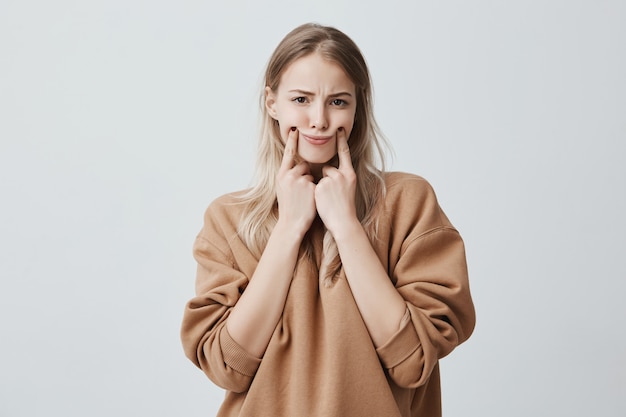 beautiful blonde woman making grimace, touching her cheeks with fingers, making herself smile, frowning face, being displeased and upset. Face expression and negative emotions