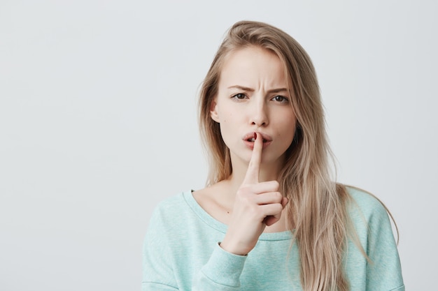 Beautiful blonde woman shows hush sign, has serious expression, asks for silence.