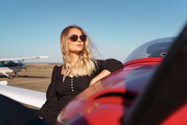 Free photo beautiful blonde woman wearing modern sunglasses posing near a red private plane with sky